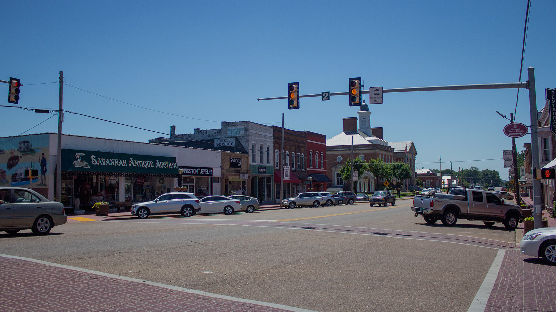 A ground view of the Hardin County Chamber of Commerce building with adjacent buildings and cars parked in front of the building. Three traffic lights can be seen with yellow lit up. The background shows a blue bright sky.
