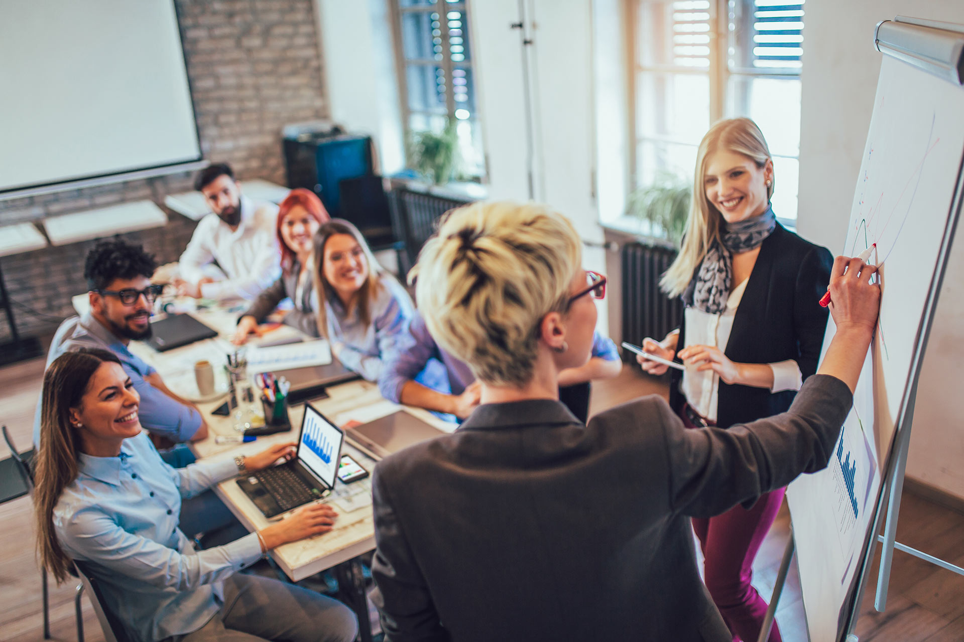 A team leader with blonde hair and glasses writes on a chart at the front of a conference room, engaging with team members seated around the table. The group, consisting of men and women, smiles while collaborating, with laptops open on the tables, fostering a productive and positive atmosphere.