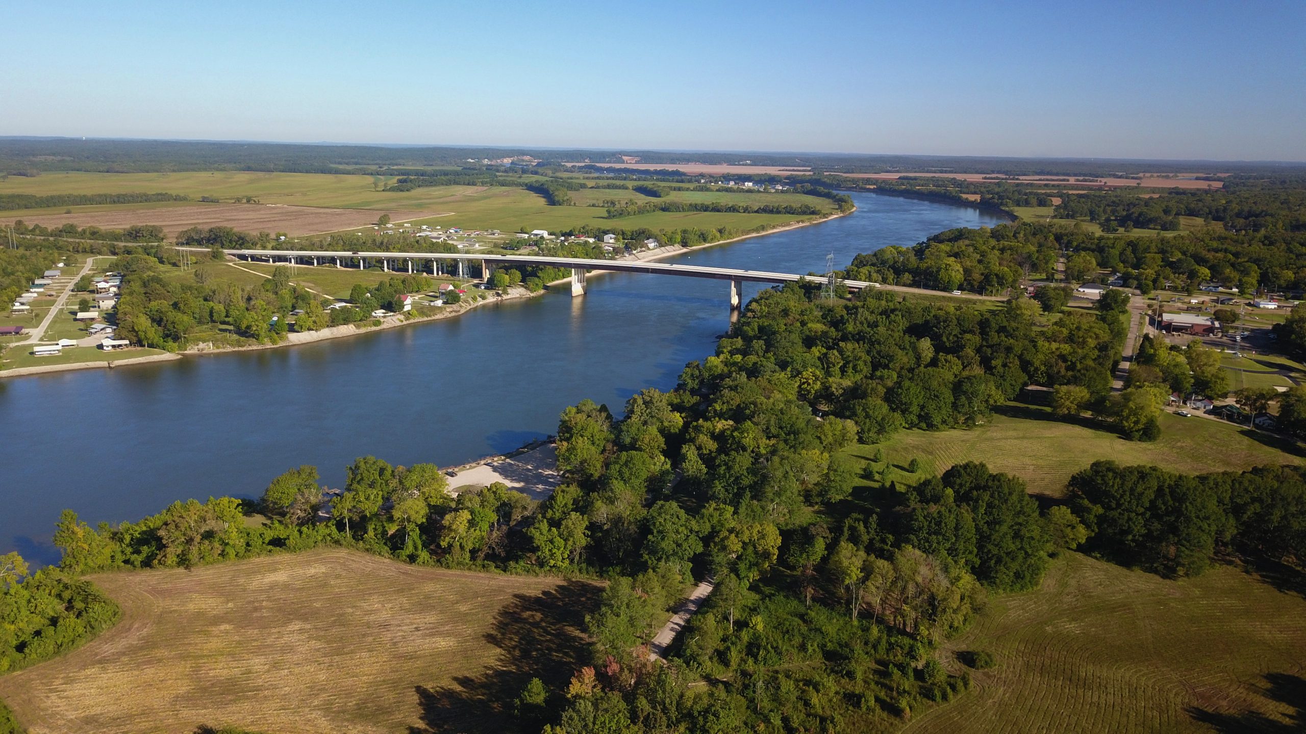 An aerial drone view of the Hardin County Bridge spanning a serene river at the center, flanked by residential houses nestled among lush trees. The scene captures a stunning blue sky and a picturesque horizon in the background, highlighting the natural beauty and charm of the area.